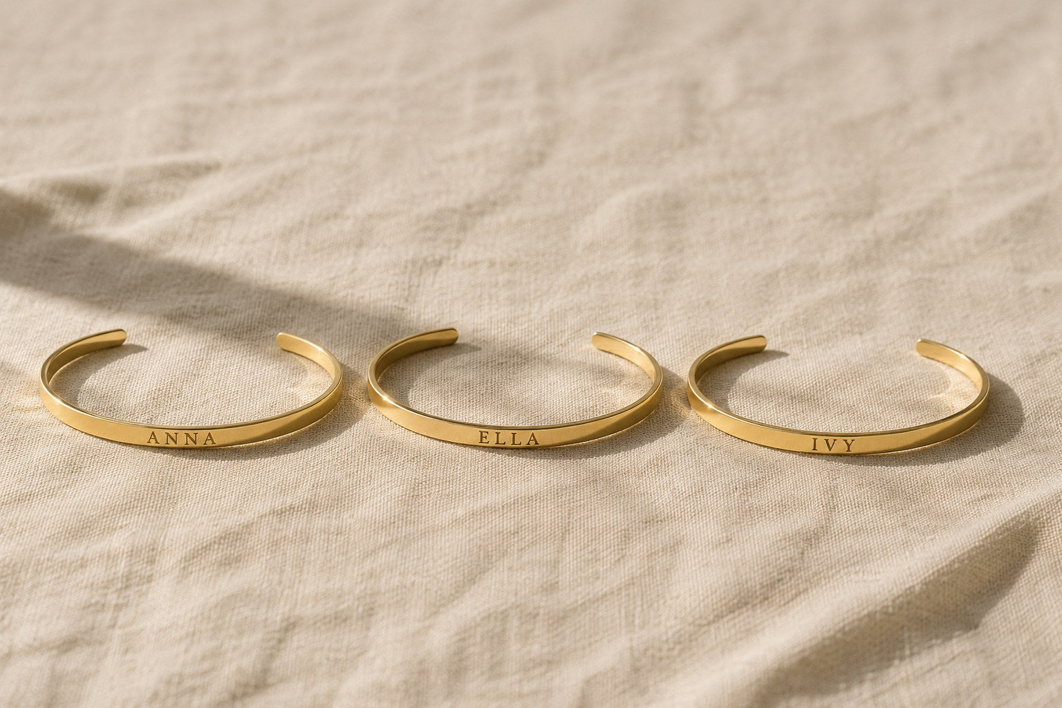 Three engraved gold cuff bracelets reading ANNA, ELLA and IVY on cream linen