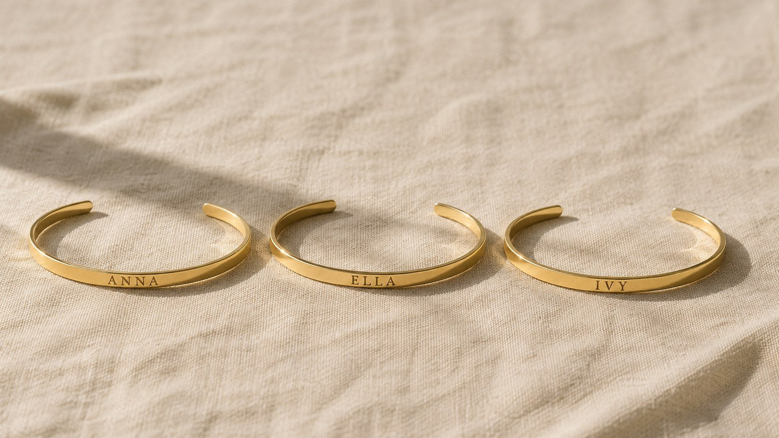 Three engraved gold cuff bracelets reading ANNA, ELLA and IVY on cream linen