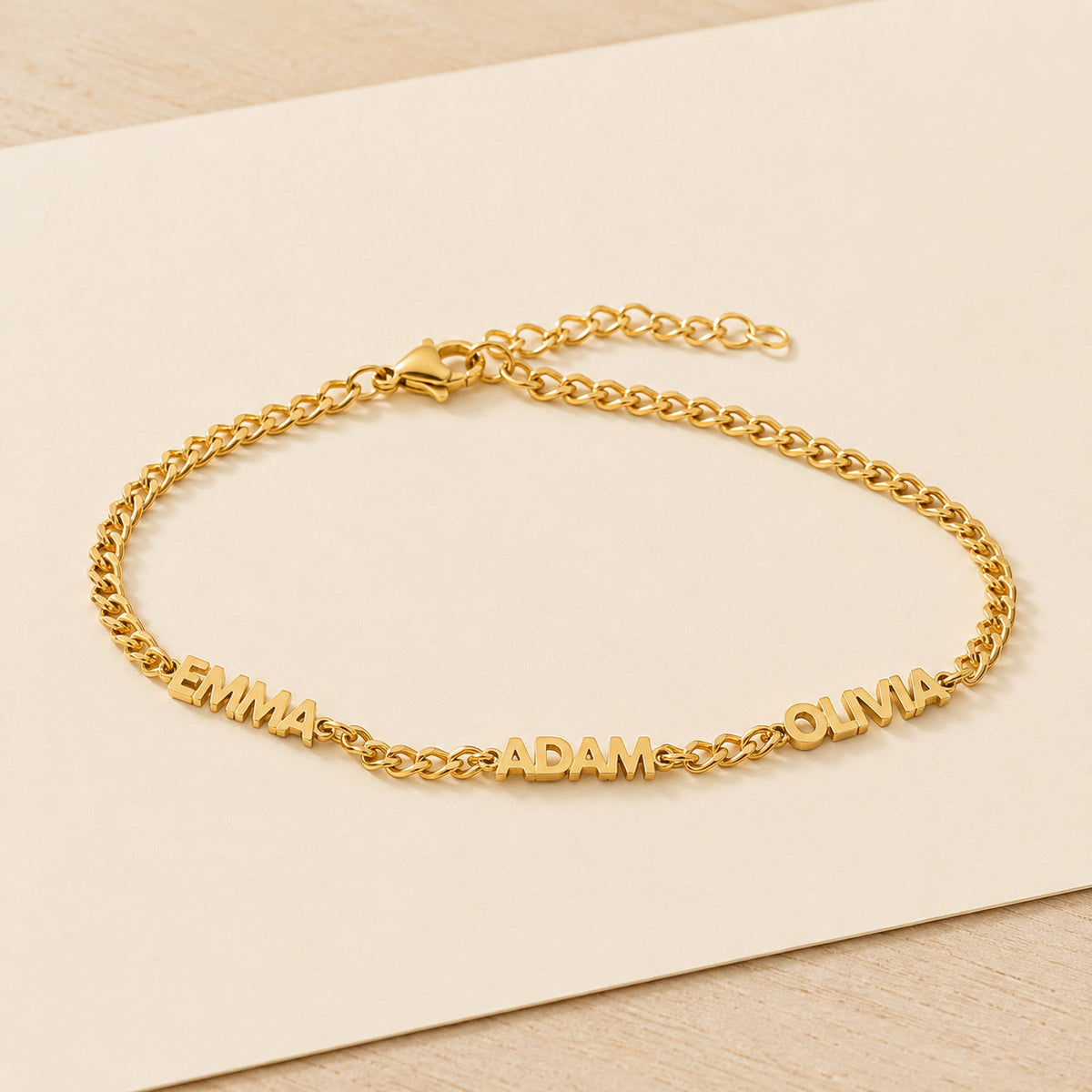 Gold name bracelet with three names resting on a blank cream card on pale wood